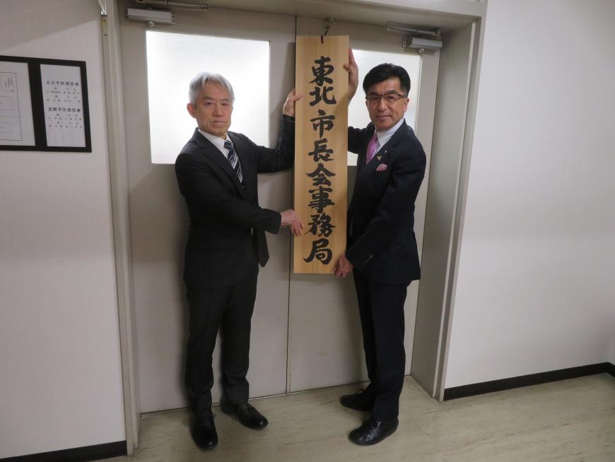 部屋の入口で「東北市長会事務局」と書かれた縦書きの看板を市長と男性で持っている写真