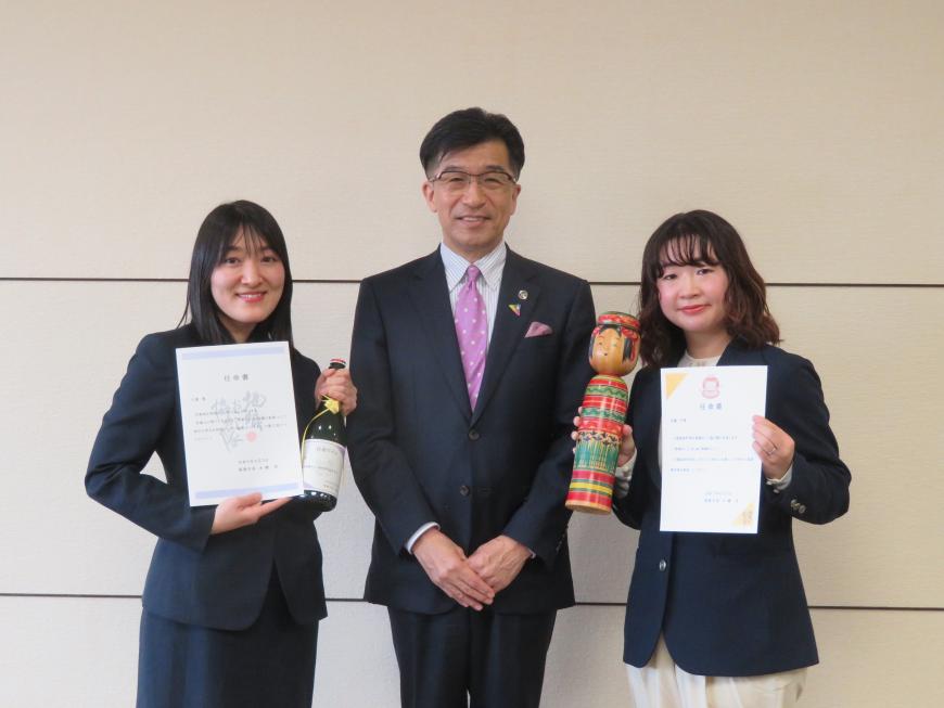市長の右隣の女性が任命書とワインボトルを持ち、左隣の女性が任命書とこけしを持っている写真