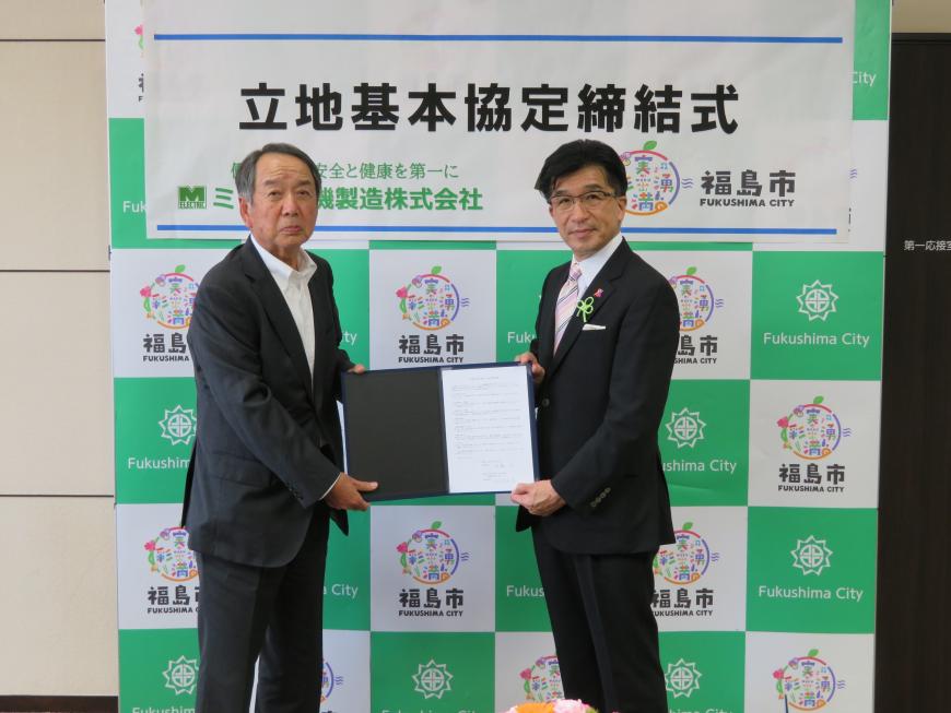 「立地基本協定締結式」と書かれた横断幕が貼られた福島市のバックボードの前で市長と男性が協定書を一緒に持ち記念撮影している写真