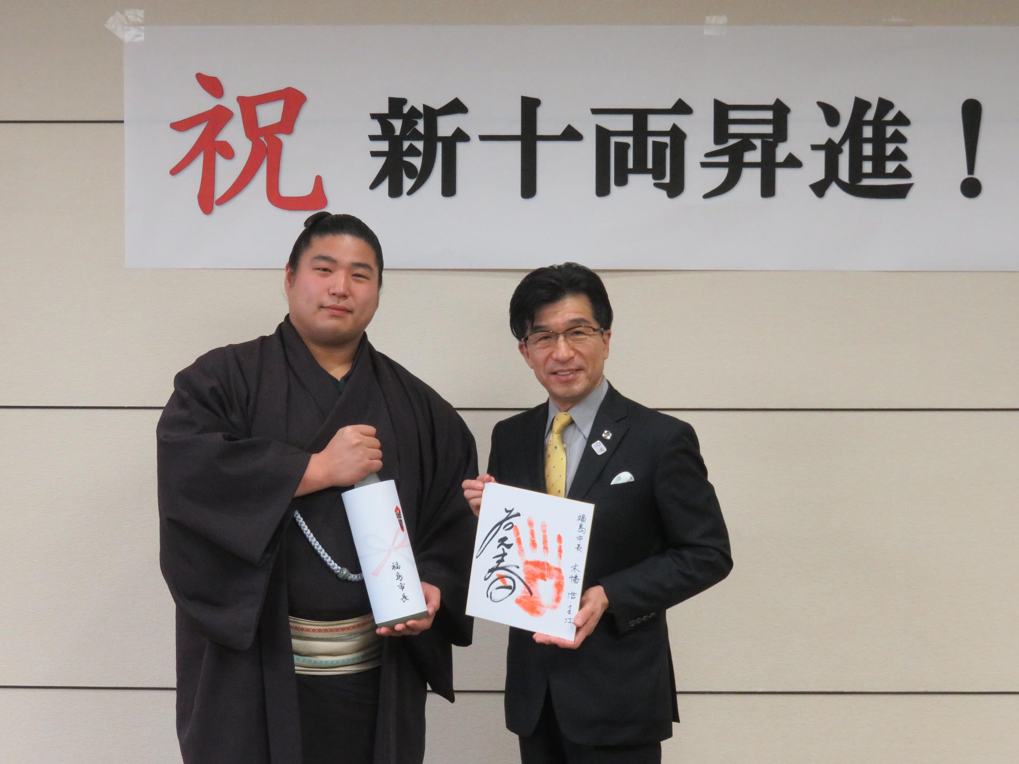 「祝 新十両昇進!」と書かれた横断幕の前でサイン入り手形を持つ市長と、熨斗紙が貼られたお酒の瓶の様な物を持つ若元春関が並んでいる記念写真