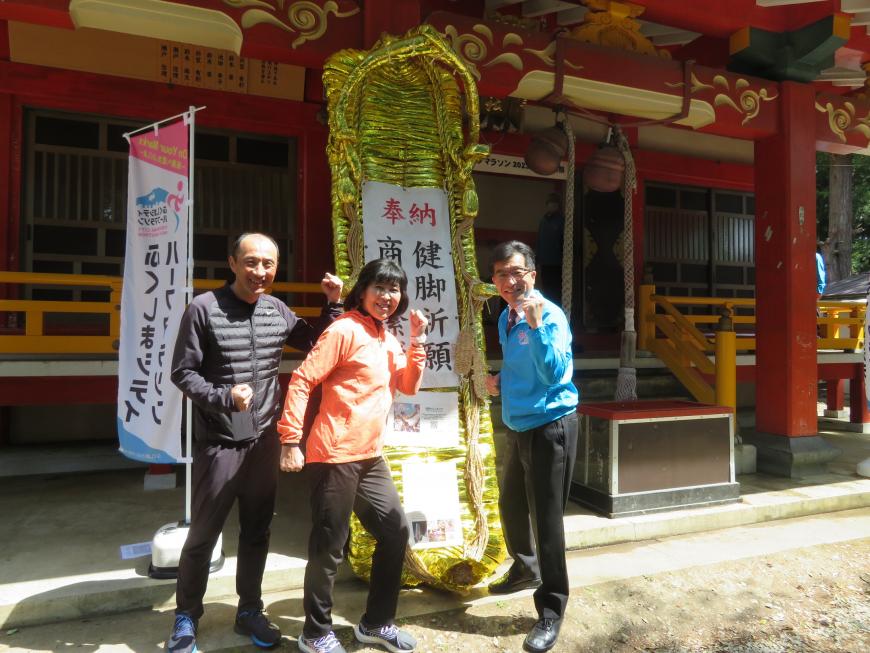 神社に奉納されている巨大な金色のわらじの前で市長と男女各1名がガッツポーズをしている写真