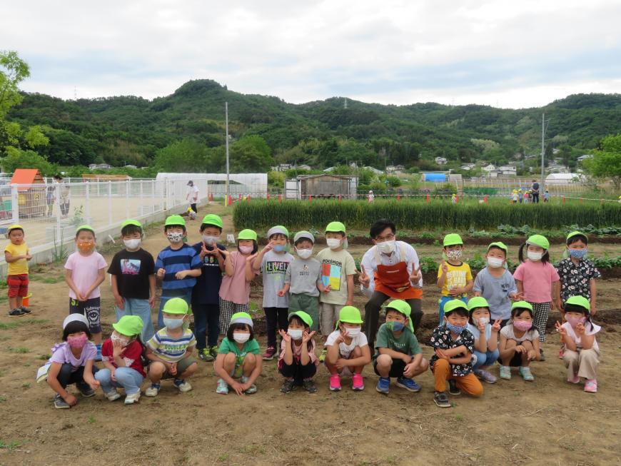 園庭前の畑で、蛍光黄緑の帽子をかぶったおかやまこども園のたくさんの園児達と市長が並んで記念撮影をしている写真