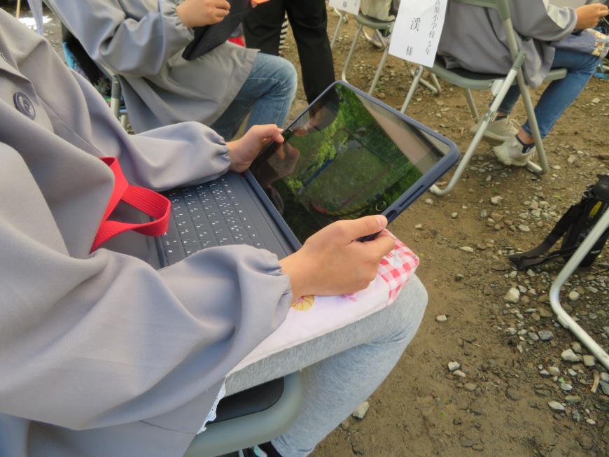 パイプ椅子に座った児童が足の上にタブレットを置き校外学習をしている写真