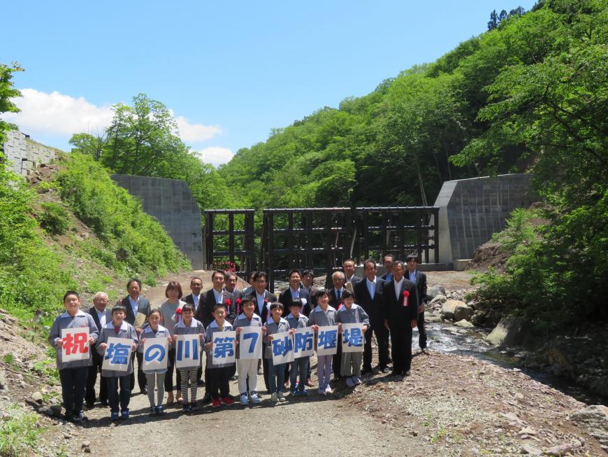 緑豊かな斜面の手前に設けられた砂防堰堤の前で「祝塩の川第7砂防堰堤」と書かれた一文字ポスターを持った児童達と関係者らが並び記念撮影している写真