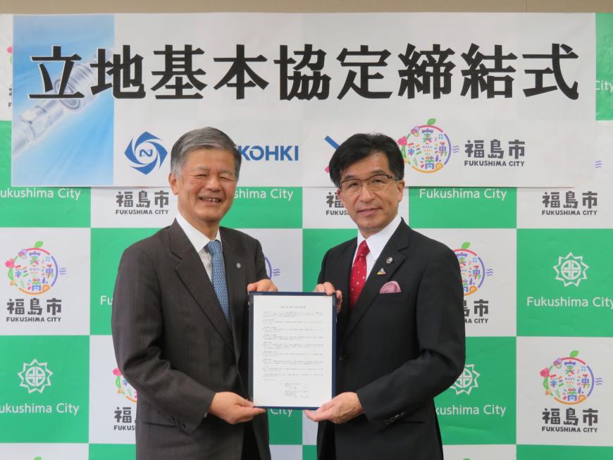 「立地基本協定締結式」の横断幕が貼ってあるバックパネルの前で、市長と男性で協定書を持っている写真
