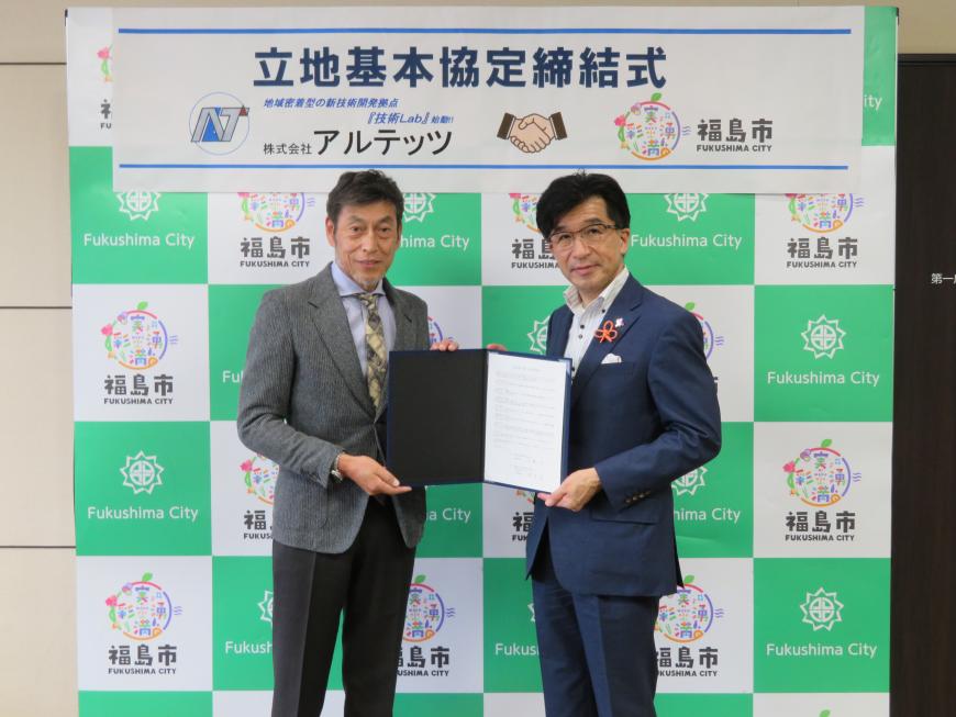 「立地基本協定締結式」と書かれた横断幕が貼られた福島市のバックボードの前で市長と男性が協定書を一緒に持ち、記念撮影している写真