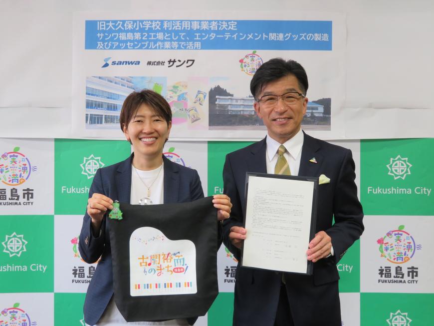 バックパネルの前で、市長が協定書を持ち、隣の女性が「古関裕而のまち」の文字がプリントされた布を持っている写真