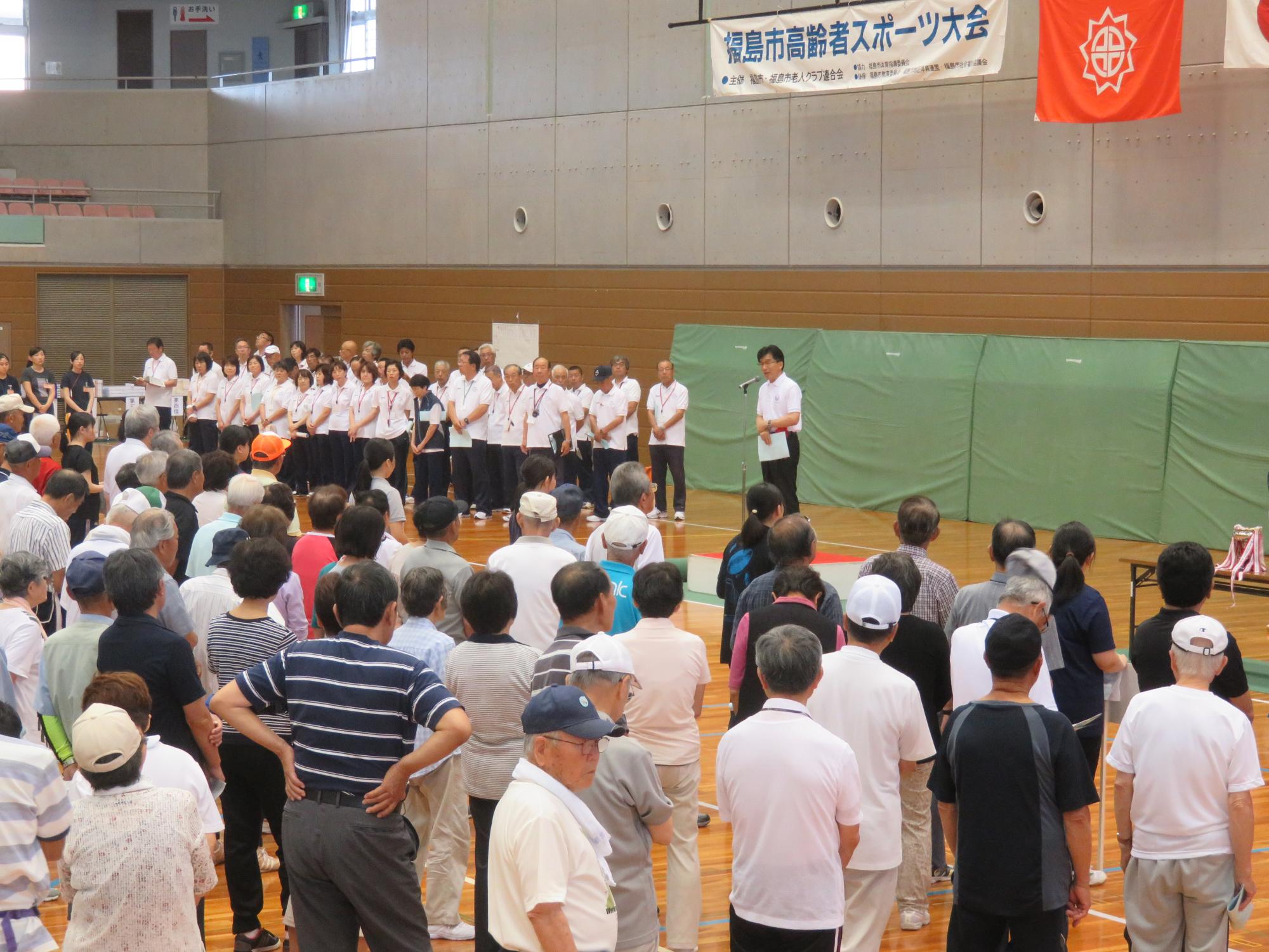 体育館にたくさんの高齢者が整列して立っており、向かいの壁側の左手に白いシャツと黒いズボンを着たたくさんの関係者たちが集まって立っている中、関係者の右側に立った市長が話をしている写真