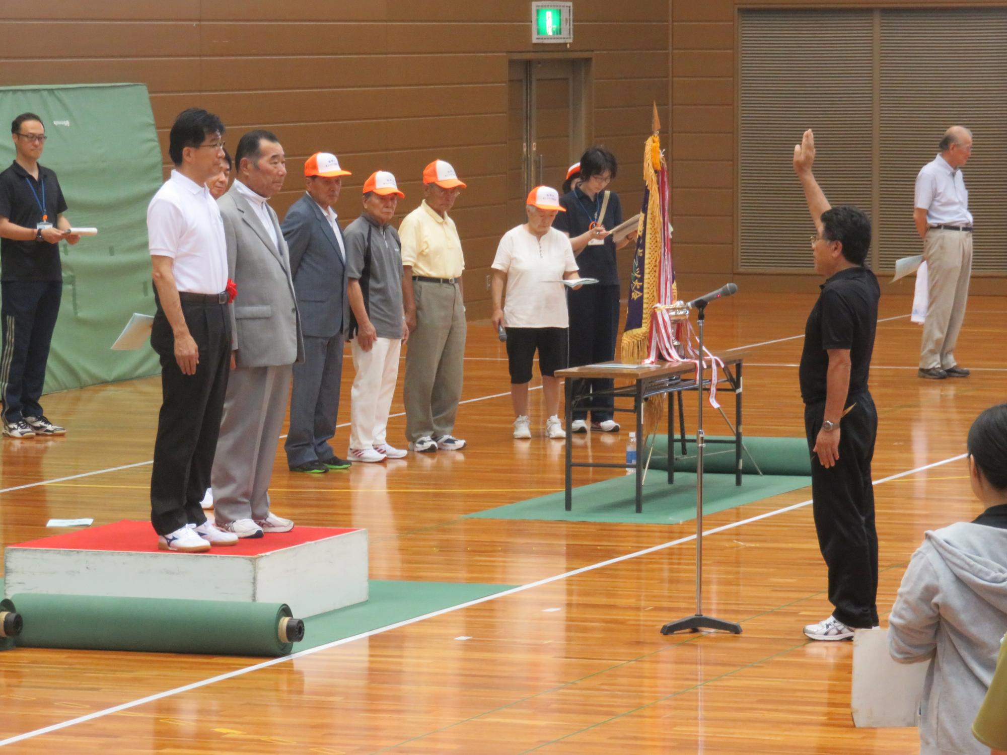 体育館で白とオレンジ色の配色の帽子をかぶった男性が4名並んで立った前に優勝旗が置かれたテーブルが設置されており、その左側の台の上に立った市長とグレーのスーツの男性に向かって、黒の上下を着た男性が右手を挙げて宣誓している様子の写真
