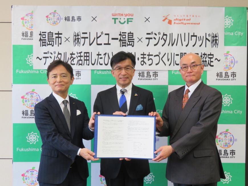 バックパネルの前で、市長を中心にして立っている男性3名で協定書を持っている写真