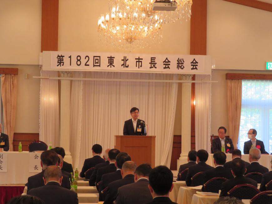 「第182回東北市長総会」の幕がある部屋で、多くの参加者の前で市長が演台に立ち、話をしている写真