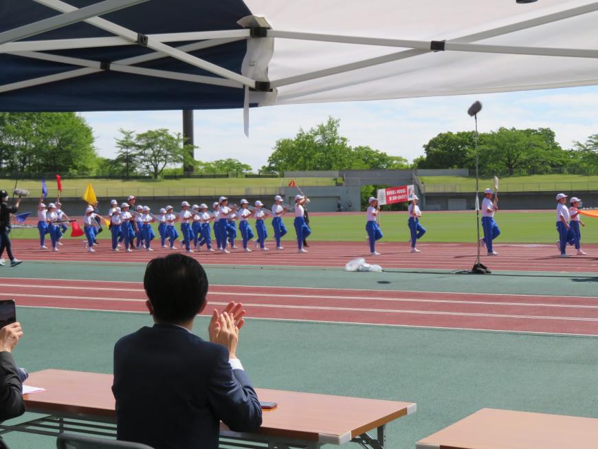 体操服を着た鼓笛隊の児童がグランドを行進しているところを、拍手をした市長が座ったテント内の来賓席から撮影した写真