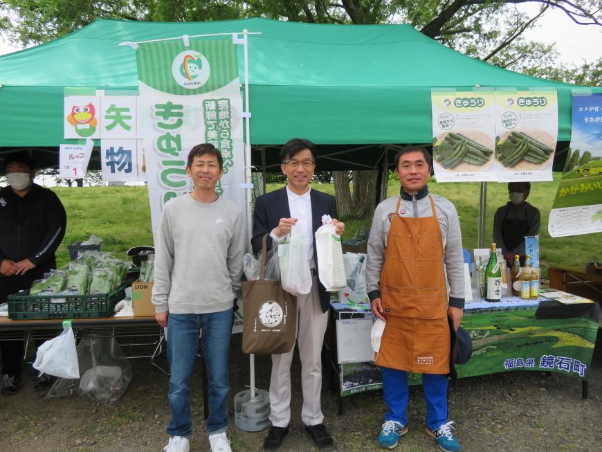 緑色のテントの前でマイバッグを下げ、ビニール袋を持った市長の両脇に男性が立っている写真