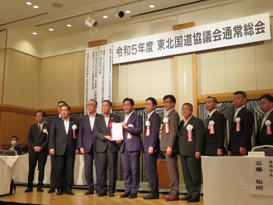 市長を含む男性12名がステージに立ち、中心に立っている2名で書類を持っている写真