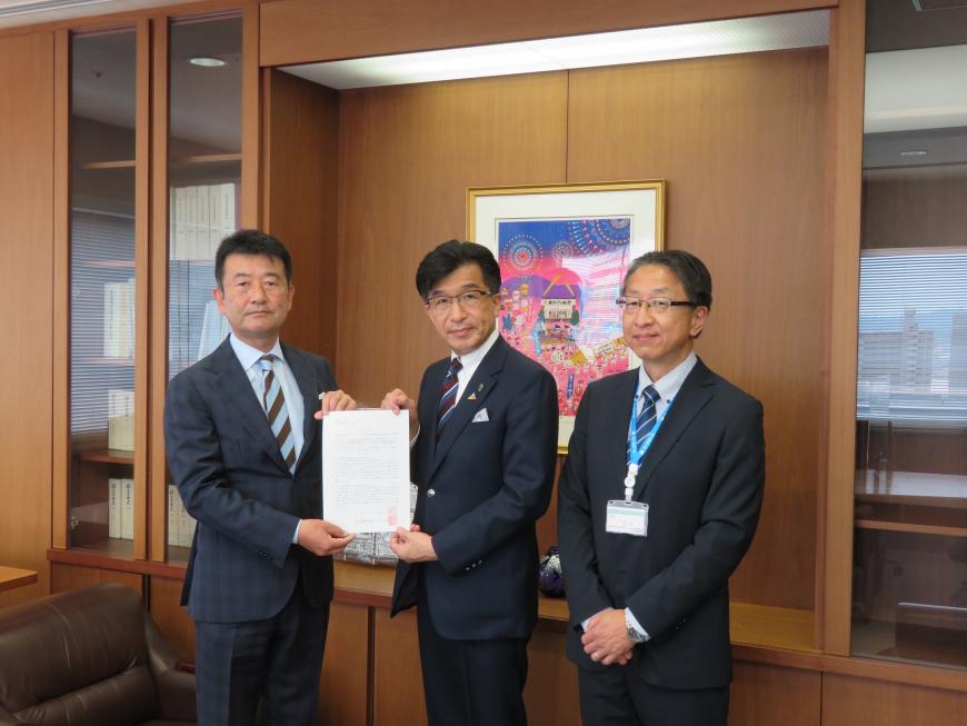 執務室内で男性と市長で要望書を持ち、その横に男性が立っている写真