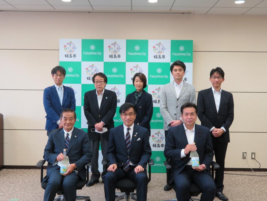 福島市のバックパネルの前で、前列の椅子に市長と瓶をを持った男性2人が座り、後列に女性1人、男性4人が立っている写真