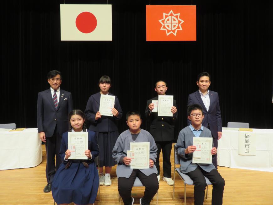 国旗と市旗が掲げられたステージ上で、表彰された児童、生徒がそれぞれ賞状を持ち、前列に3人が座り、後列に2人が市長と男性の間に立っている写真