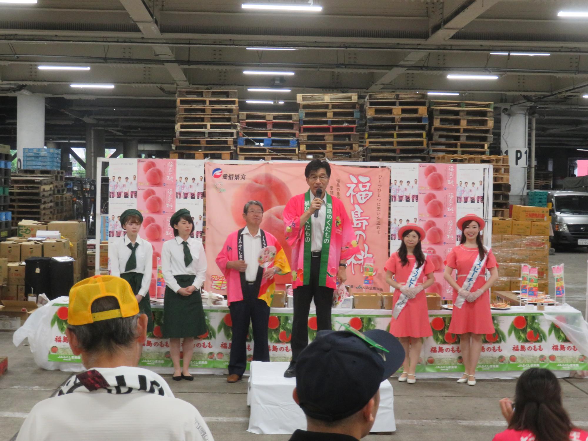 市場の中で、大きな桃のポスターの前に緑色の帽子、ネクタイ、スカートに白いシャツを着た女性2名、ピンク色の法被を着た男性、ピンク色の帽子とワンピースを身に着けた女性2名が1列に並んで立ち、その前に置かれた台に立ったピンク色の法被を着た市長がマイクを持って関係者に向かって話をしている写真