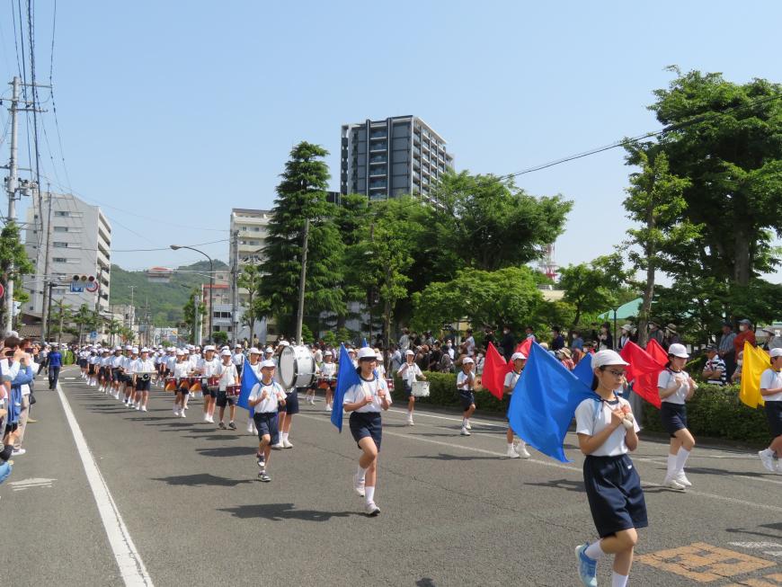 青空の下、沿道で多くの人が見ている中、旗を両肩に担いでパレードしている児童たちの後方には鼓笛隊が見えている写真