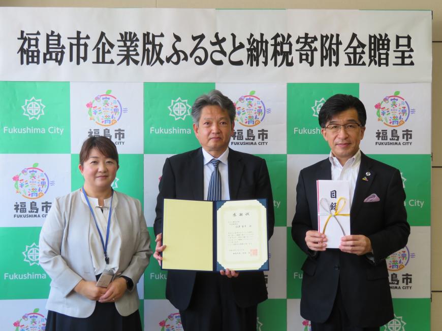 バックパネルの前で、市長が目録封筒を持ち、男性が感謝状を持ち、その横に女性が立っている写真