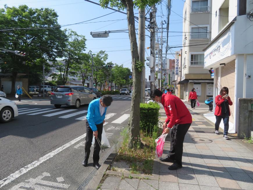 青と赤の服を着た男性2名がビニール袋を持ち、街路樹回りのゴミを拾っている写真