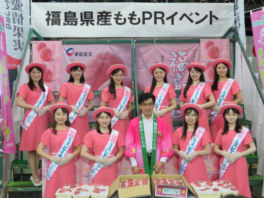 「福島県産ももPRイベント」と書かれた横断幕の前にピンク色の帽子とワンピースを着た10人の女性たちが2段になって並び、前列の中央にピンク色の法被を着た市長が立って、手前に並んだたくさんの桃一緒に写した記念写真
