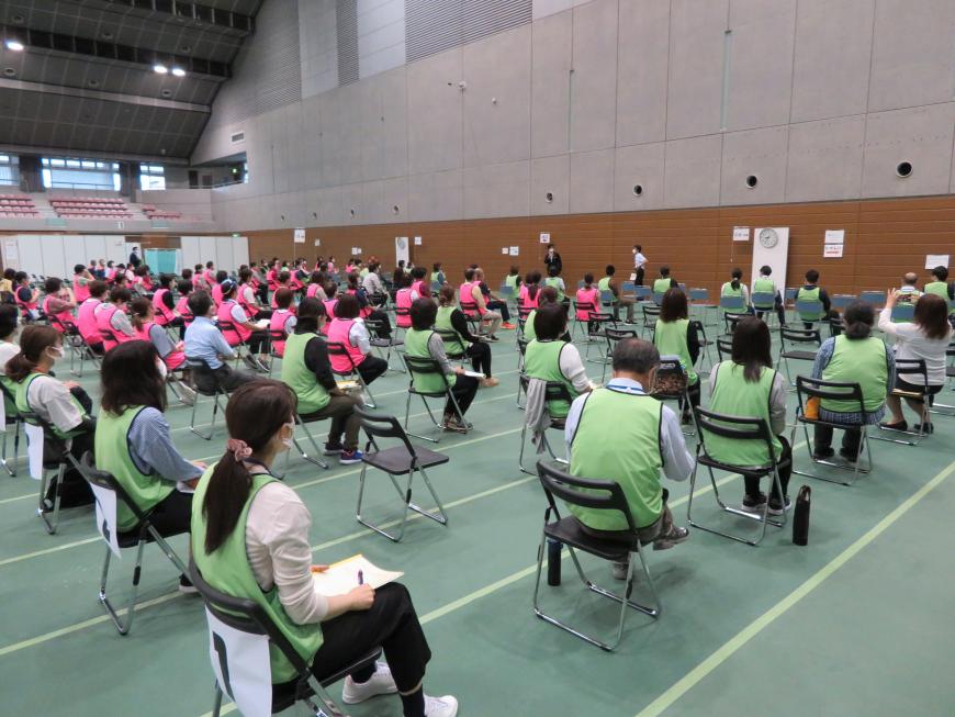 広い体育館の床が緑色の線で仕切られており、何列にも並んだたくさんの椅子に緑やピンクのビブスをつけた人々が座っている写真