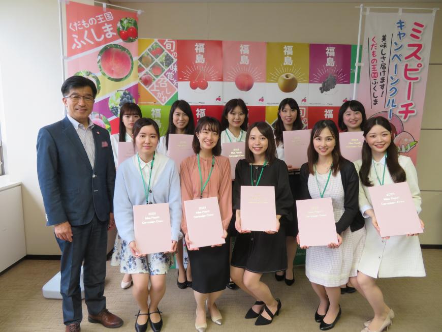 ミスピーチキャンペーンクルーに選ばれた10名の女性が果物が描かれた幟を背景に市長の横に2列に並んでいる写真