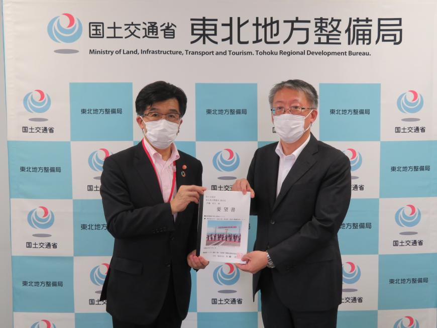 「国土交通省 東北地方整備局」と書かれたバックボードの前で市長と男性が要望書に手を携えて立っている記念写真