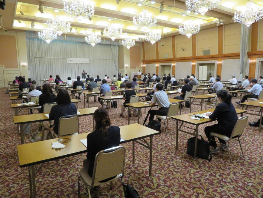 天井にシャンデリアが並んでいる広い会場に長机が間隔をあけて並んでおり、奥のステージの方を向いてたくさんの出席者が席に座っている会場全体の様子の写真