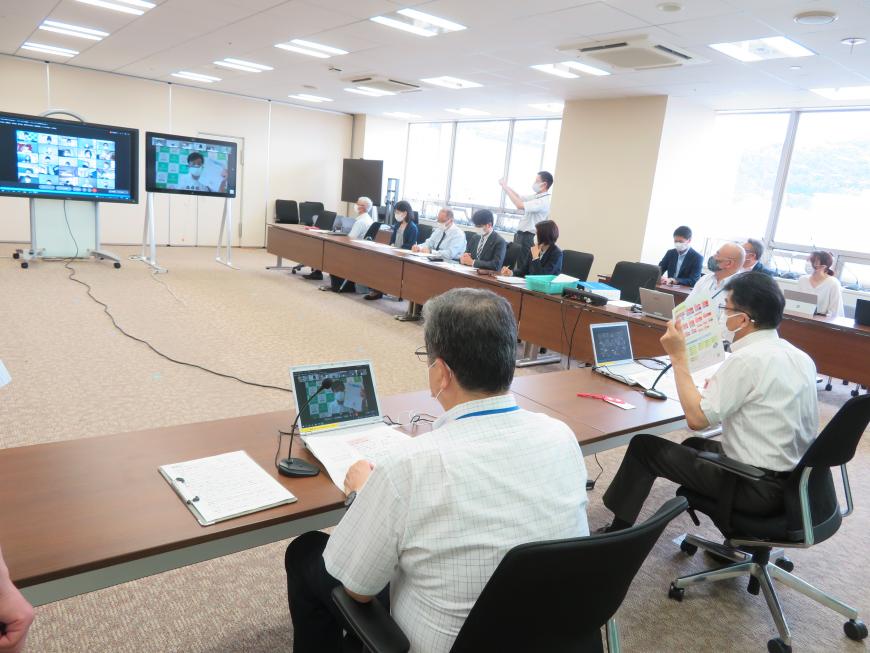 広い会議室にL字型に置かれた会議机の席に座った出席者たちが中央に設置された大きな2つのモニターを見ている様子の写真