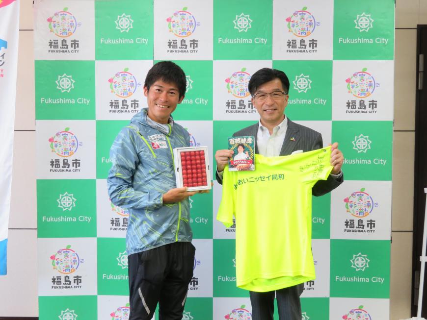 バックパネルの前で、市長が黄色のTシャツを広げて持ち、川内選手が箱詰めのさくらんぼを持って立っている写真