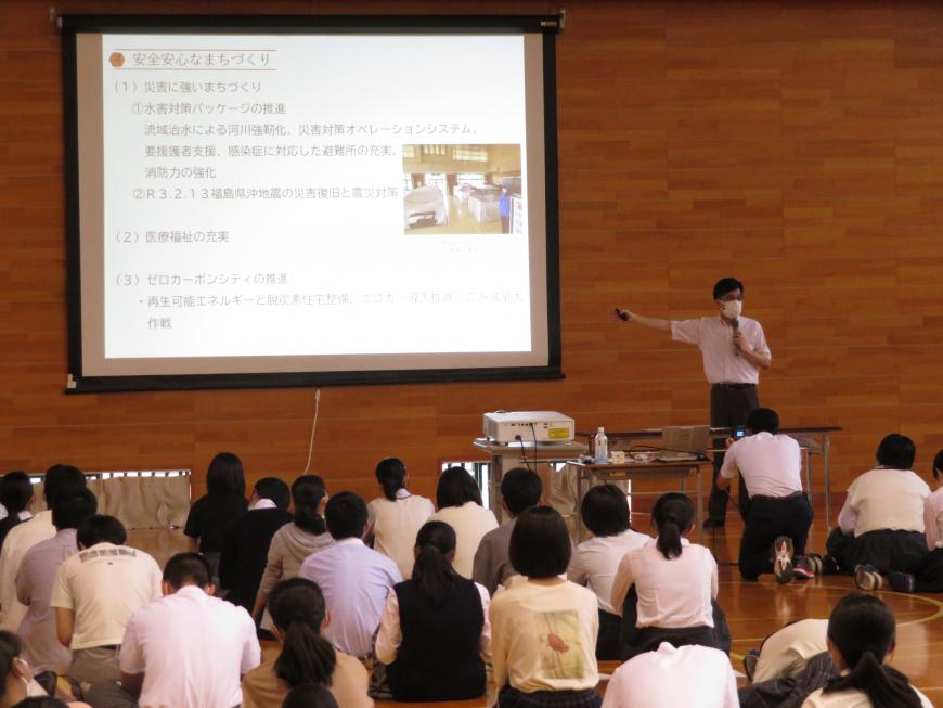 体育館の壁に設置された大きなスクリーンにスライドが映し出され、その右側に立ってマイクを持って話をしている市長と床に座って話を聞いている生徒たちの写真