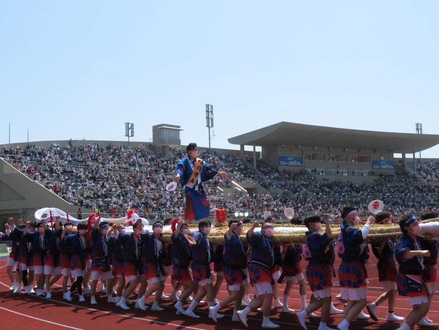 青空の下、スタジアムで多くの人が担いでいる巨大わらじに市長が乗り、立っている写真