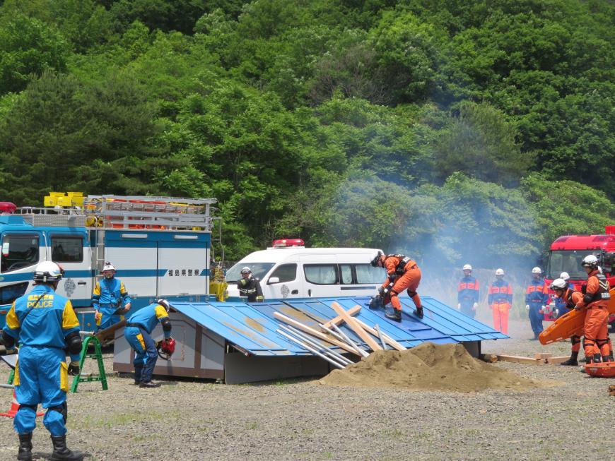 訓練している広場で青やオレンジの作業服を着た警察官や消防隊員が他の隊員が見ている中、チェンソーで屋根や壁を切っている写真