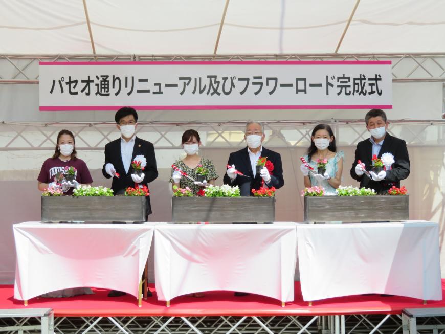 「パセオ通りリニューアル及びフラワーロード完成式」と書かれた横断幕が掲げられたテントに設置された白いクロスがかかったテーブルに置かれた3つの寄せ植えの前で、市長と男女5名がそれぞれ寄せ植えに花を植えるポーズをとって写した記念写真