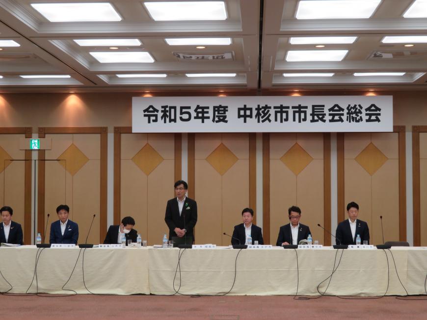 「令和5年度中核市市長会総会」のボードが吊るされた会場で、横一列に座っている参加者の中で市長が自席で立って話をしている写真