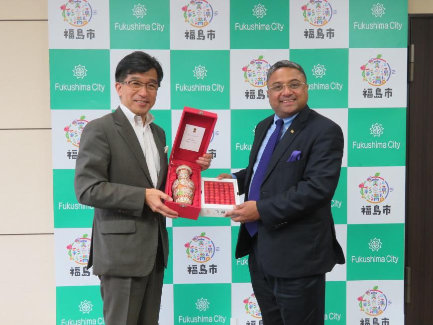 バックパネルの前で市長が箱に入った壺を持ち、横の男性が箱詰めのさくらんぼを持っている写真
