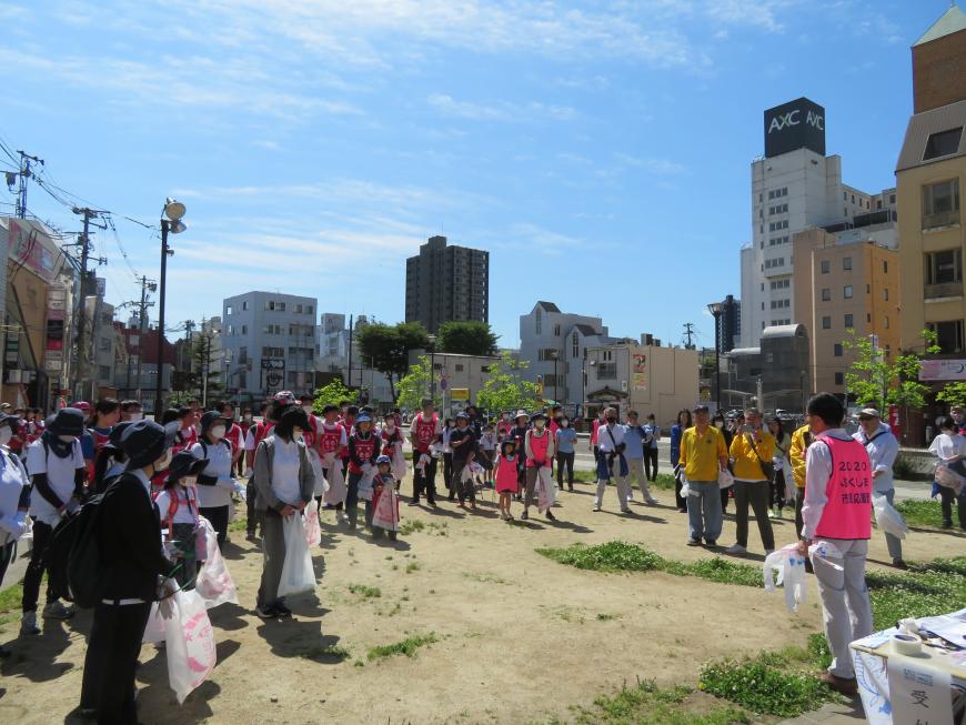 青空の下、それぞれビニール袋を持った参加者が集まっている前で、市長が話をしている写真