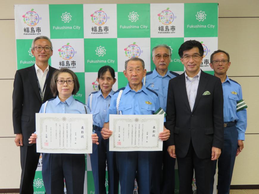 福島市のバックボードの前で水色のシャツと紺色のズボンを着用した5人の男女と市長、関係者の男性1名が並んで立ち、前列の男女がそれぞれ賞状を持って写った記念写真