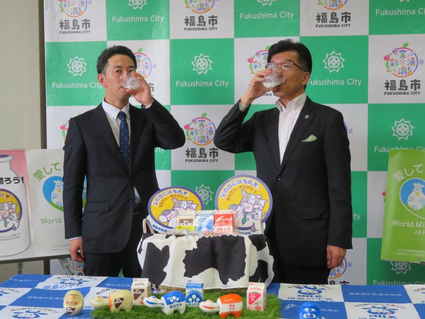 バックパネルの前で市長と男性がコップに入った牛乳を飲んでいる写真
