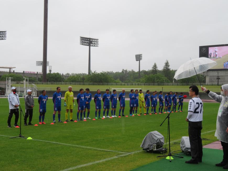 雨の中、芝生のグラウンドに整列している青色や黄色のユニフォーム姿の選手に向けて、白いユニフォームを着た市長が挨拶をしている写真