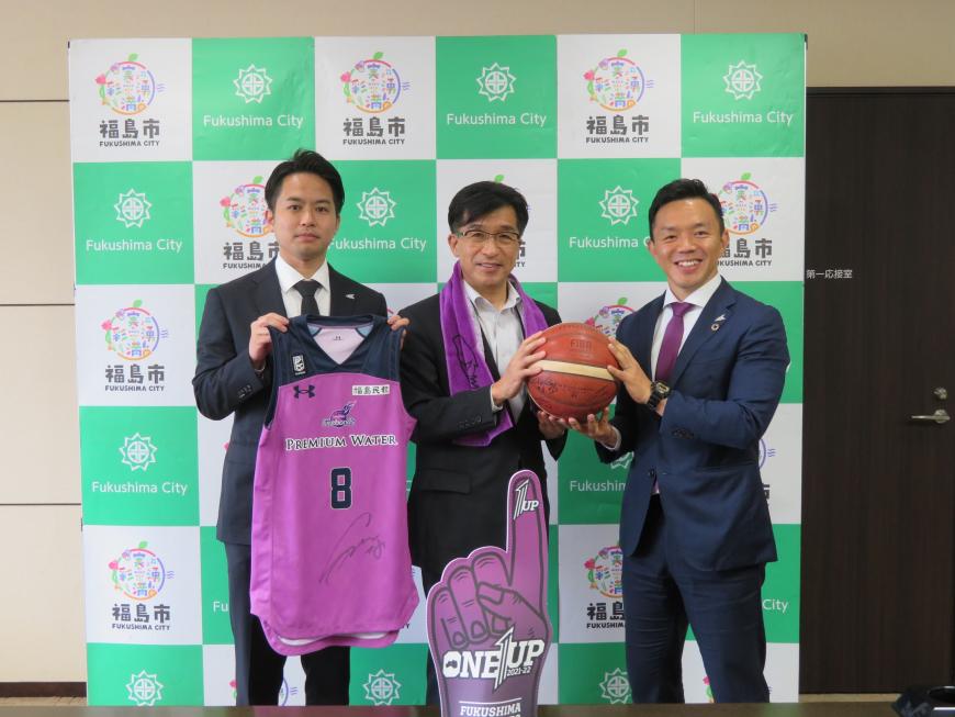 福島市のバックパネルの前で、紫色のタオルを首からかけた市長が右隣の男性と一緒にバスケットボールを持ち、左隣の男性が紫色のユニフォームを持って並んで立った記念写真