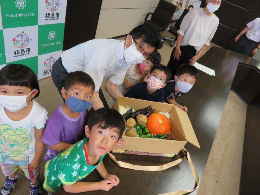 長机の上にジャガイモ、ナス、ピーマン、カボチャなどの野菜が詰まった段ボール箱が置いてあり、それを腰をかがめて手に取っている市長と市長を囲んで立っている小さな子供たちがこちらを見ている写真