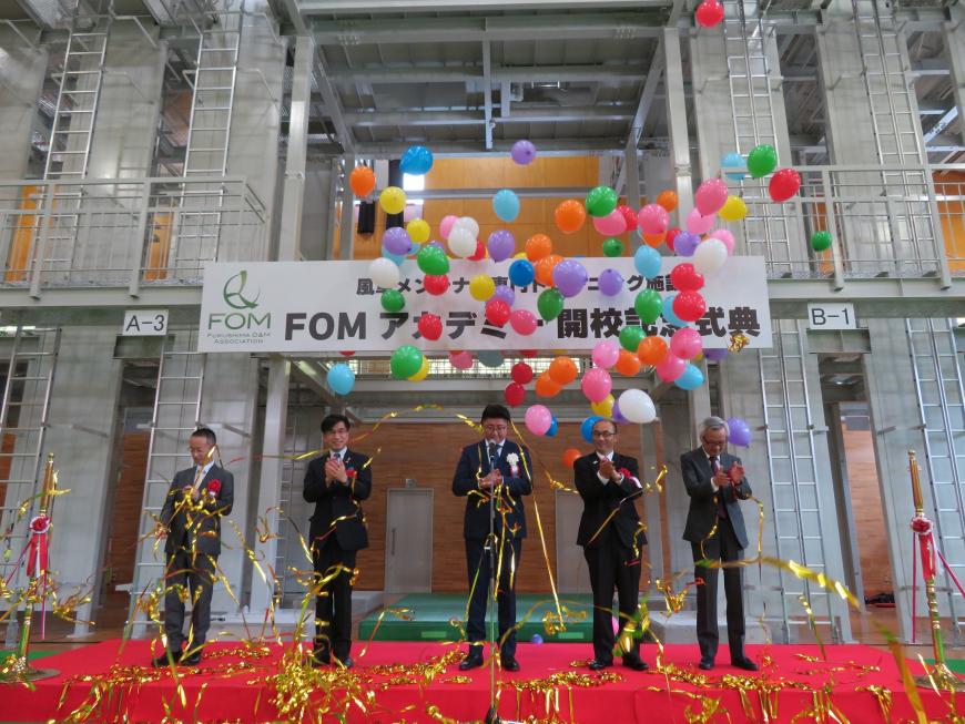 「FOMアカデミー開校記念式典」と書かれた横断幕が飾られた会場で、色とりどりの風船が飛び、金色の紙テープが舞っている中、赤いカーペットが敷かれた舞台に立った市長と4人の男性たちが拍手をしている写真