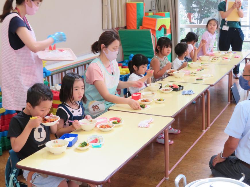 こども園の教室に並んだテーブルの上に給食が配膳されており、席に着いた園児たちと保育士の女性が食事をしている様子の写真