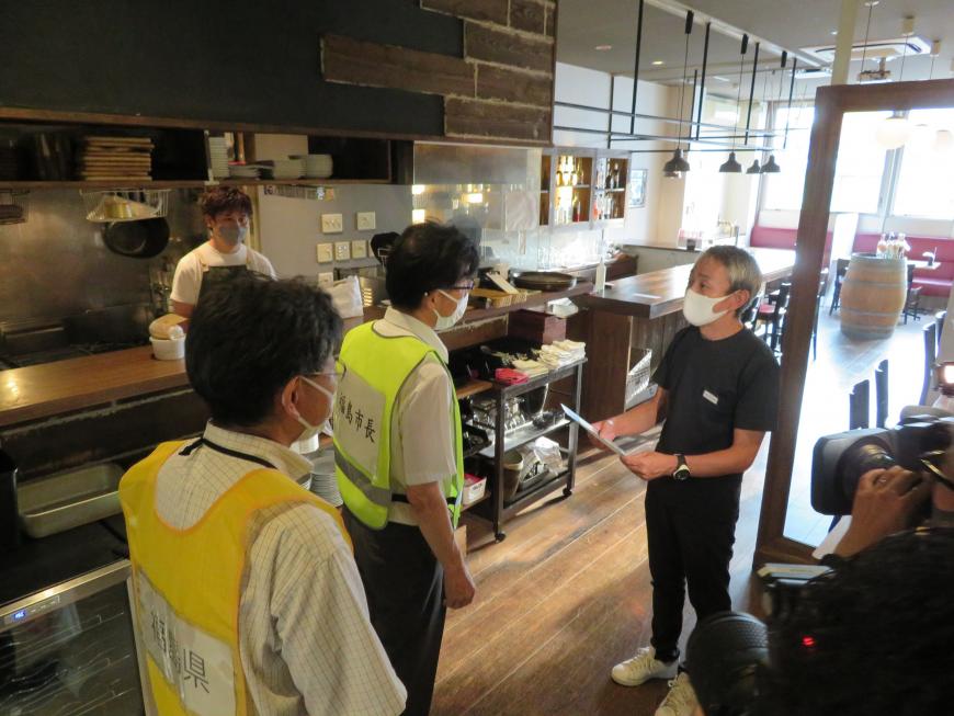 飲食店の厨房のカウンター前で黄色のビブスを着た市長が黒いTシャツとズボンを着た男性と話をしている様子の写真