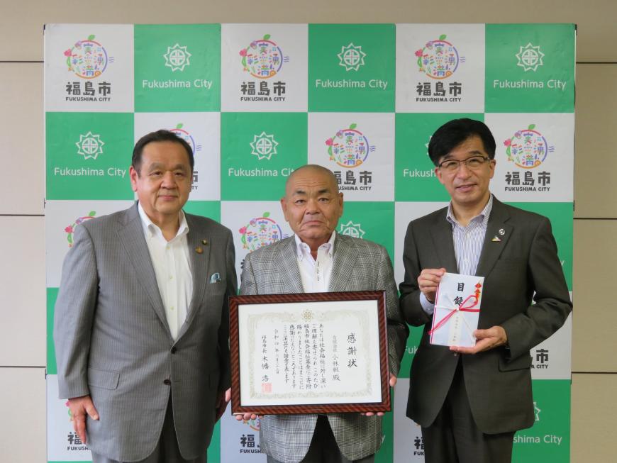 福島市のバックパネルの前で、額に入った感謝状を持った男性の両側に目録封筒を持った市長と男性1人が立っている写真