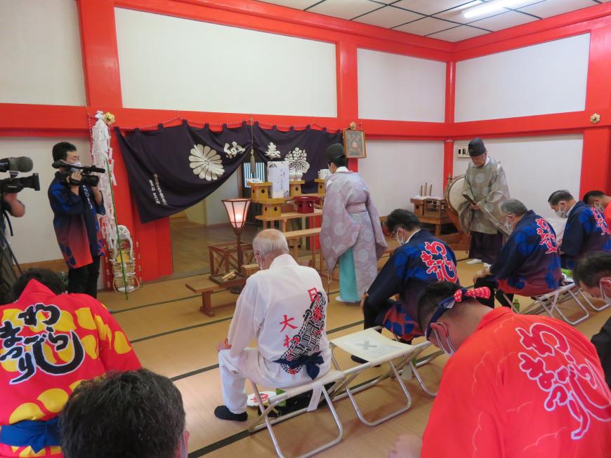 神棚へ祈祷をする宮司の後ろで、席に座った背中に「わらじまつり」と書かれた赤や紺の半被を着た方々が、頭を下げて疫病退散祈願を行っている様子の写真