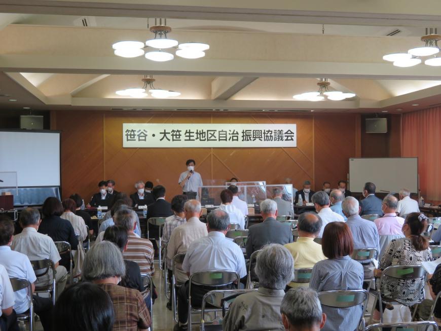 「笹谷・大笹生地区自治振興競技会」と書かれた横断幕が設置された壁の前にマイクを持って立った市長が、対面に座った大勢の出席者たちに話をしている写真